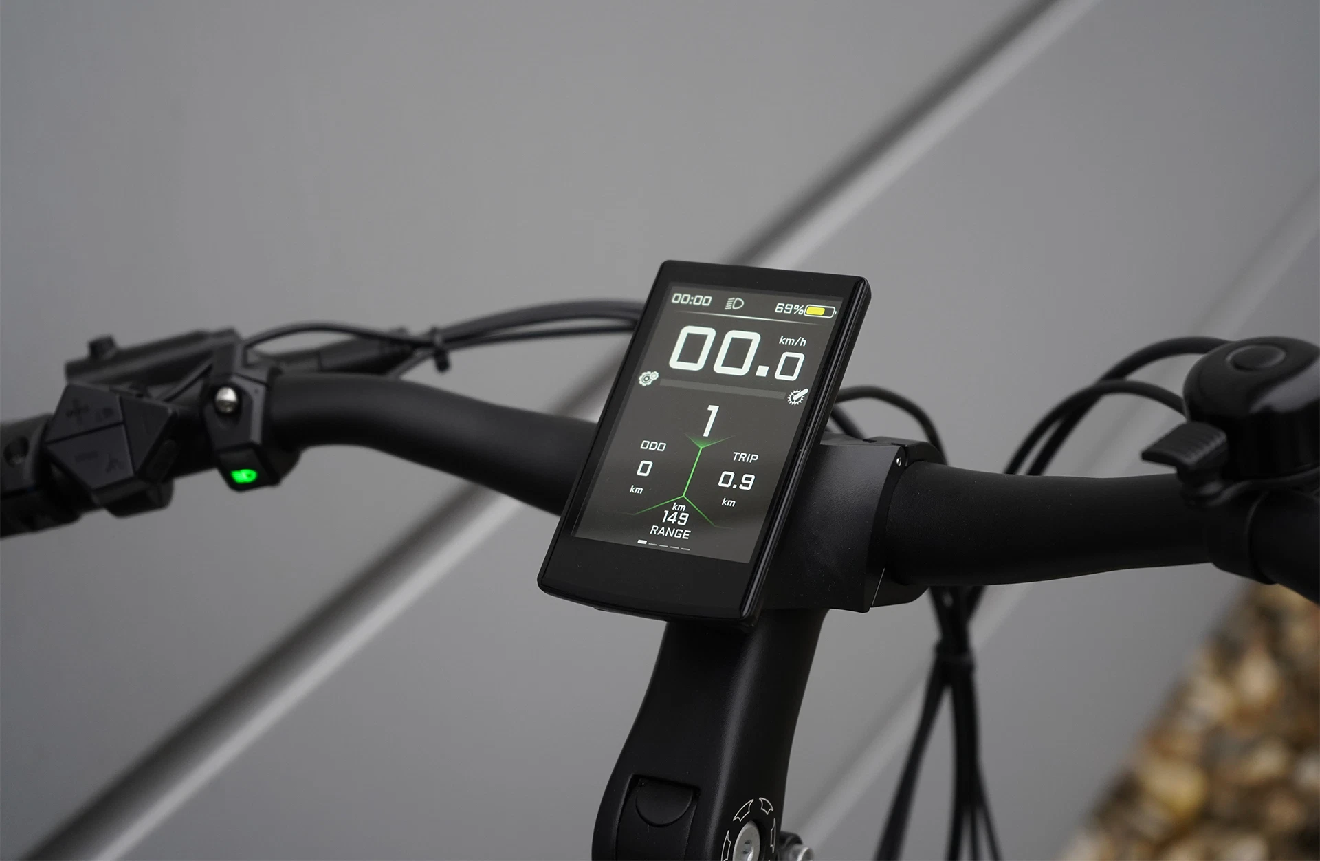 Bildausschnitt Lenker mit zentralem, vollfarbigem TFT-Display, linker Bedieneinheit samt grünem Licht und ergonomischen Griffen vor Metallwand und steinigem Untergrund