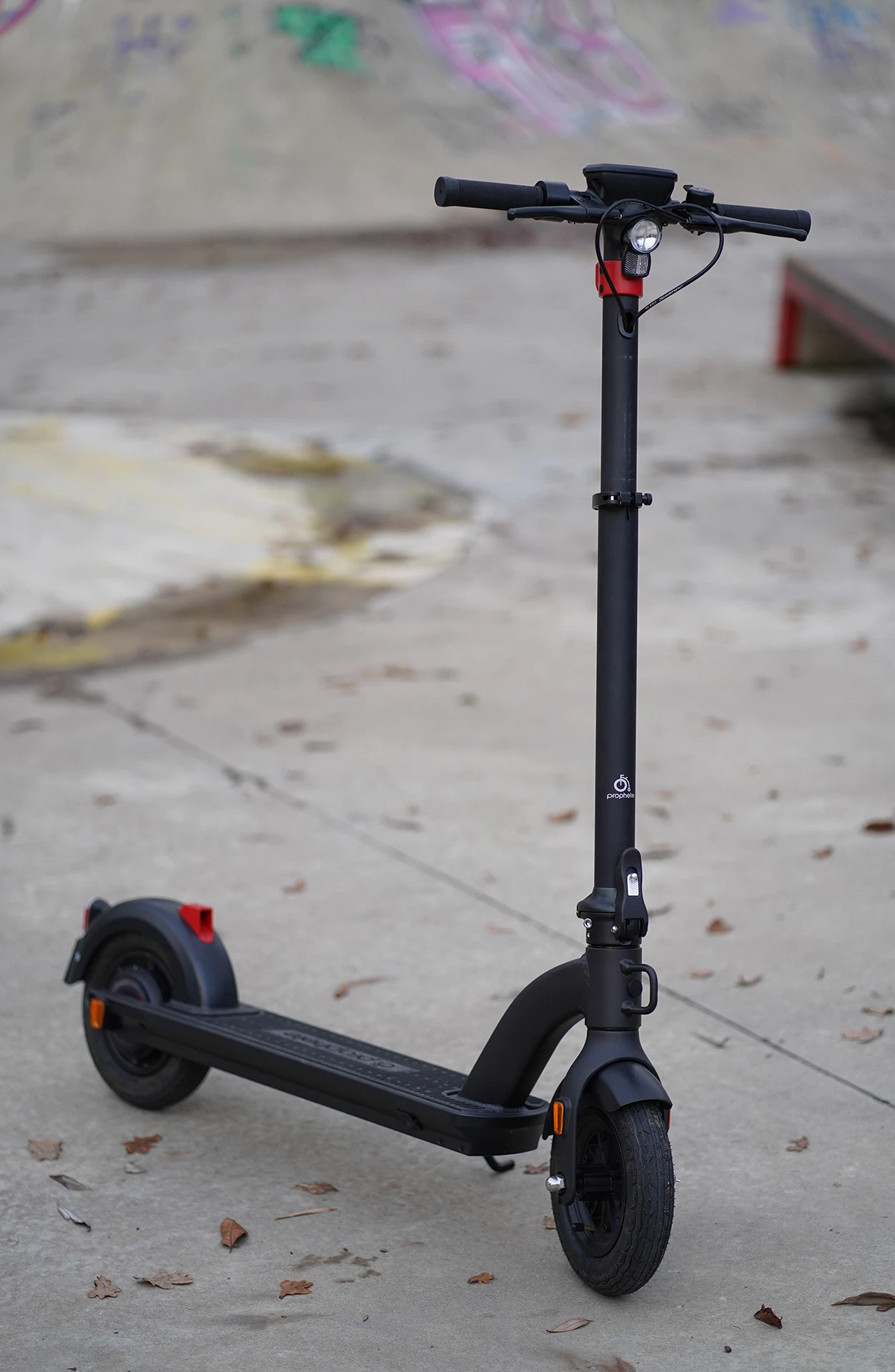 Vollansicht diagonal von vorne Prophete E-Scooter in Skateanlage mit asphaltiertem Untergrund und Blättern