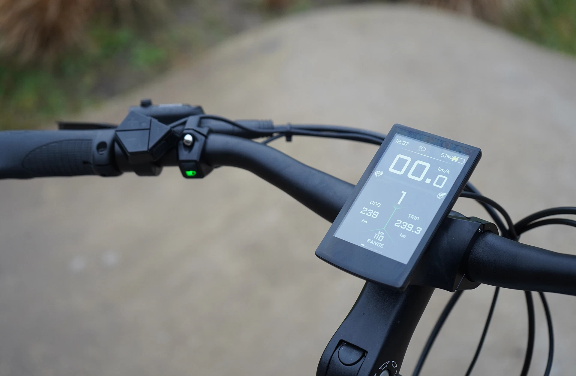 Bildausschnitt Lenkerstange mit linksseitigem Bedienelement, grünem LED-Knopf und zentralem, vollfarbigem TFT-Display auf Schotterweg.