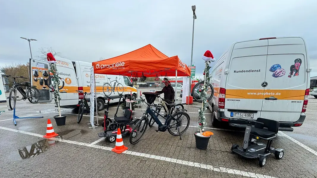 Prophete Wintercheck-Stand mit orangefarbenem Pavillon und zwei Servicefahrzeugen, E-Bikes und Scooter auf einem Parkplatz, weihnachtlich dekoriert mit Santa-Mützen.
