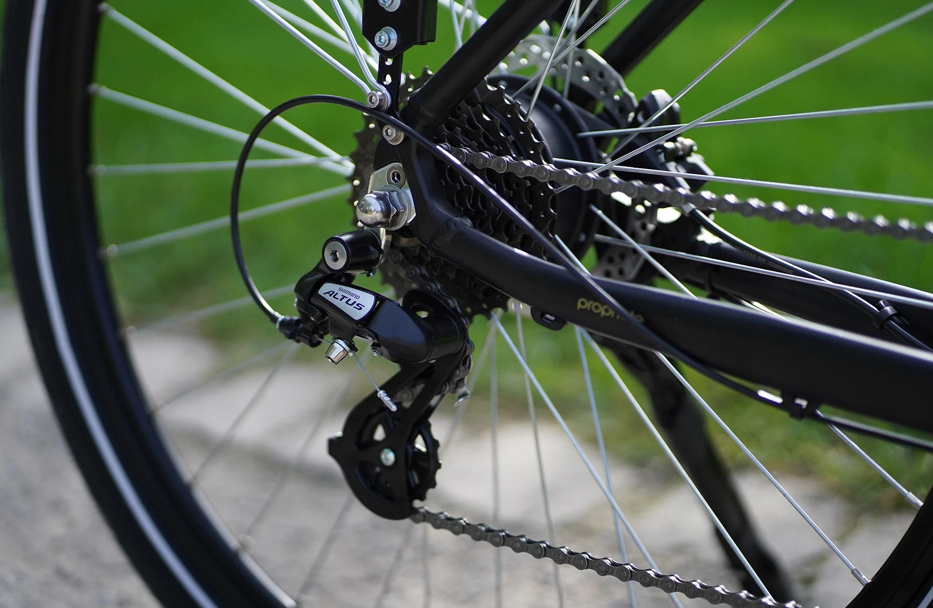 Bildausschnitt Detailansicht Shimano Altus Schaltwerk mit Kassette und Scheibenbremse, sowie Speichen und Seitenständer auf unscharfem Schotterweg und Rasen