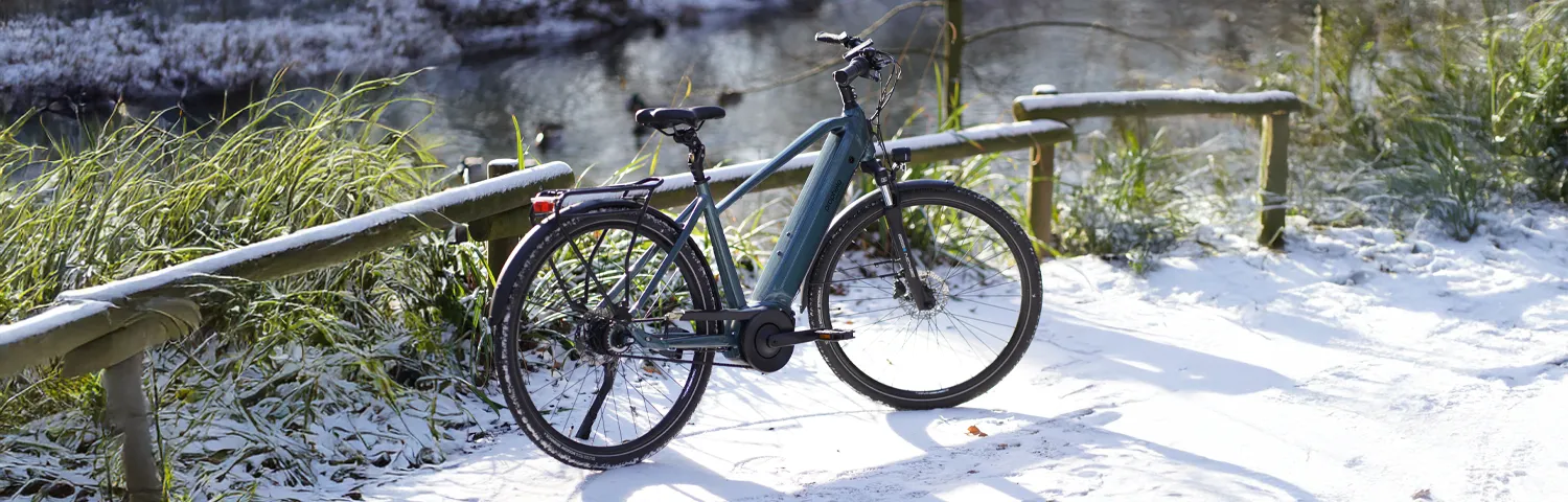 Wintercycling - Prophete Geniesser 6.0 E-Bike auf verschneitem Weg am Fluss, mit Gepäckträger und integriertem Akku.