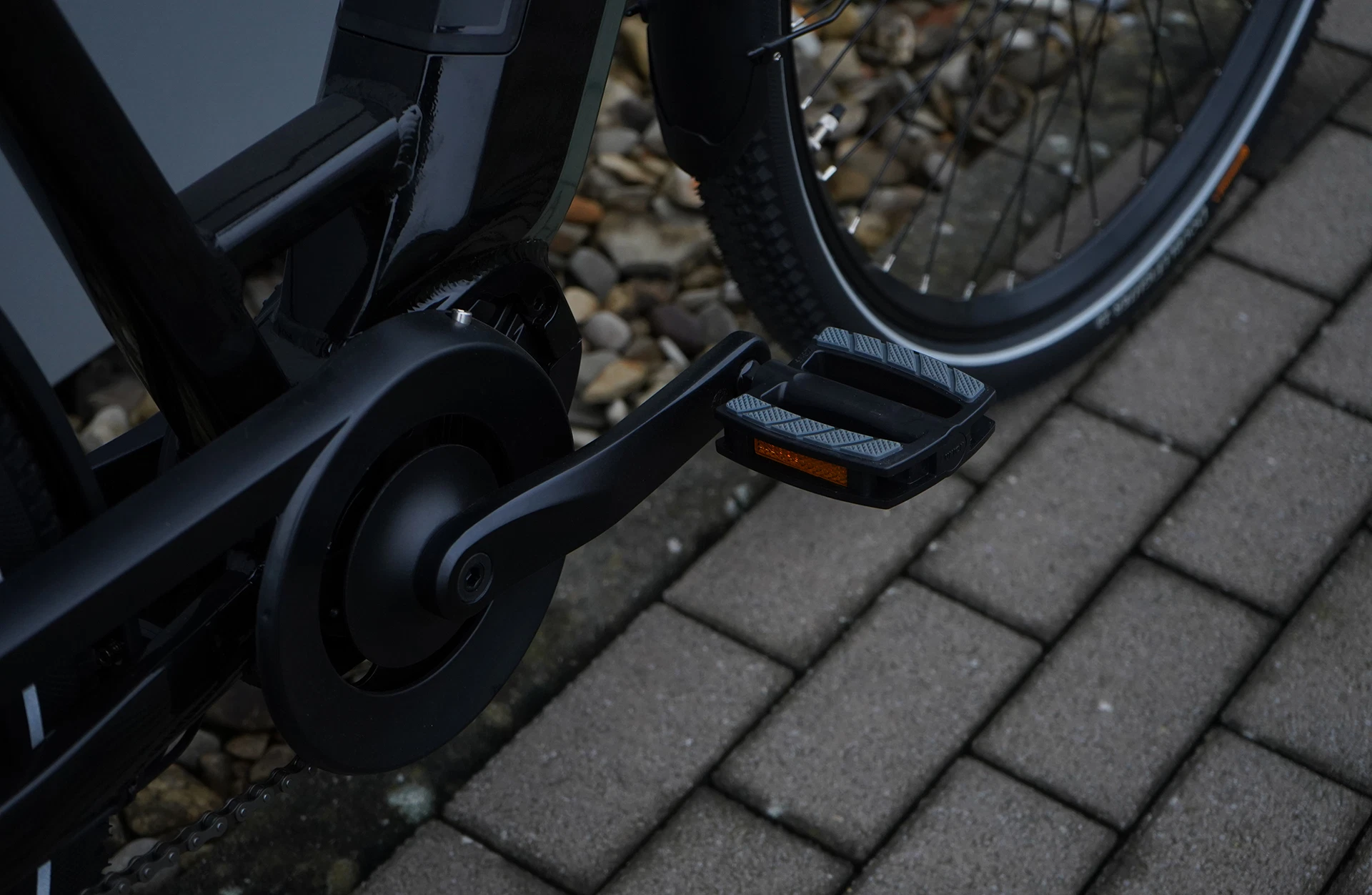 Bildausschnitt Pedal mit Reflektor und geschütztem Kettenantrieb, sowie angeschnittenem Reifen auf steinigem Untergrund