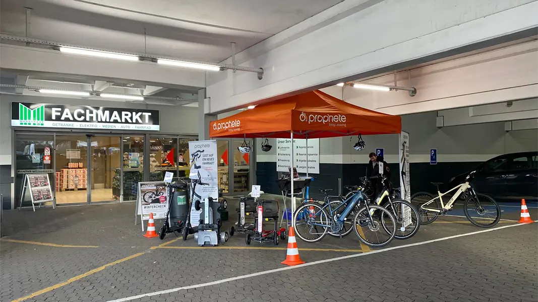 Prophete Wintercheck-Stand mit orangefarbenem Pavillon in einem überdachten Bereich vor einem Marktkauf, mehrere E-Bikes und Scooter zur Ansicht, Service-Mitarbeiter vor Ort.
