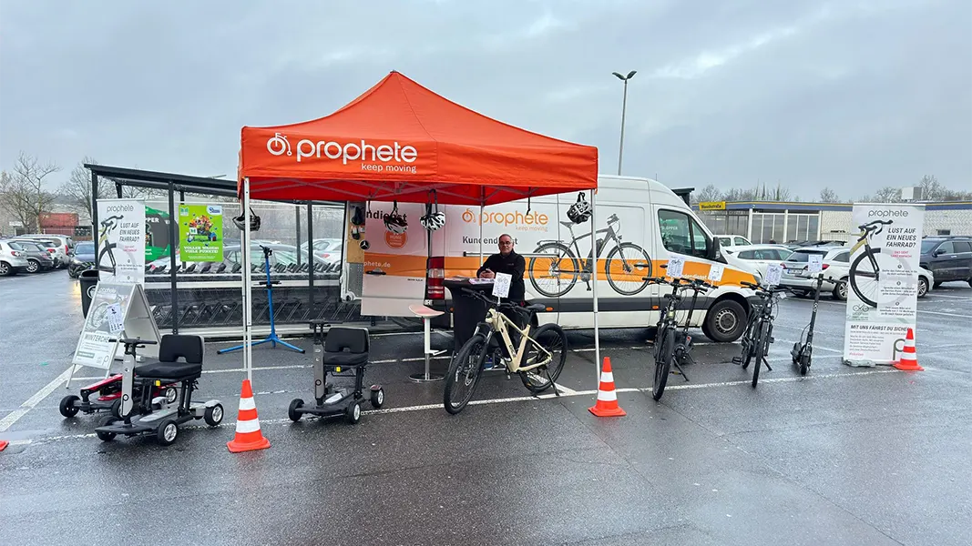 Prophete Wintercheck-Stand mit orangefarbenem Pavillon, Service-Mitarbeiter und E-Bikes vor Prophete-Servicefahrzeug auf einem Parkplatz.