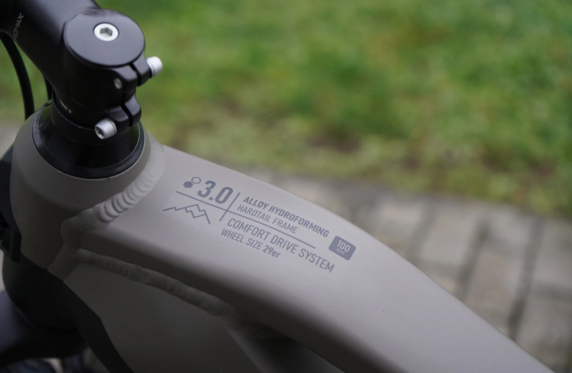 Bildausschnitt Oberrohr Prophete E-MTB DICE mit "3.0 Alloy hydroforming hardtail Frame" Beschriftung vor gepflastertem Untergrund und Rasen