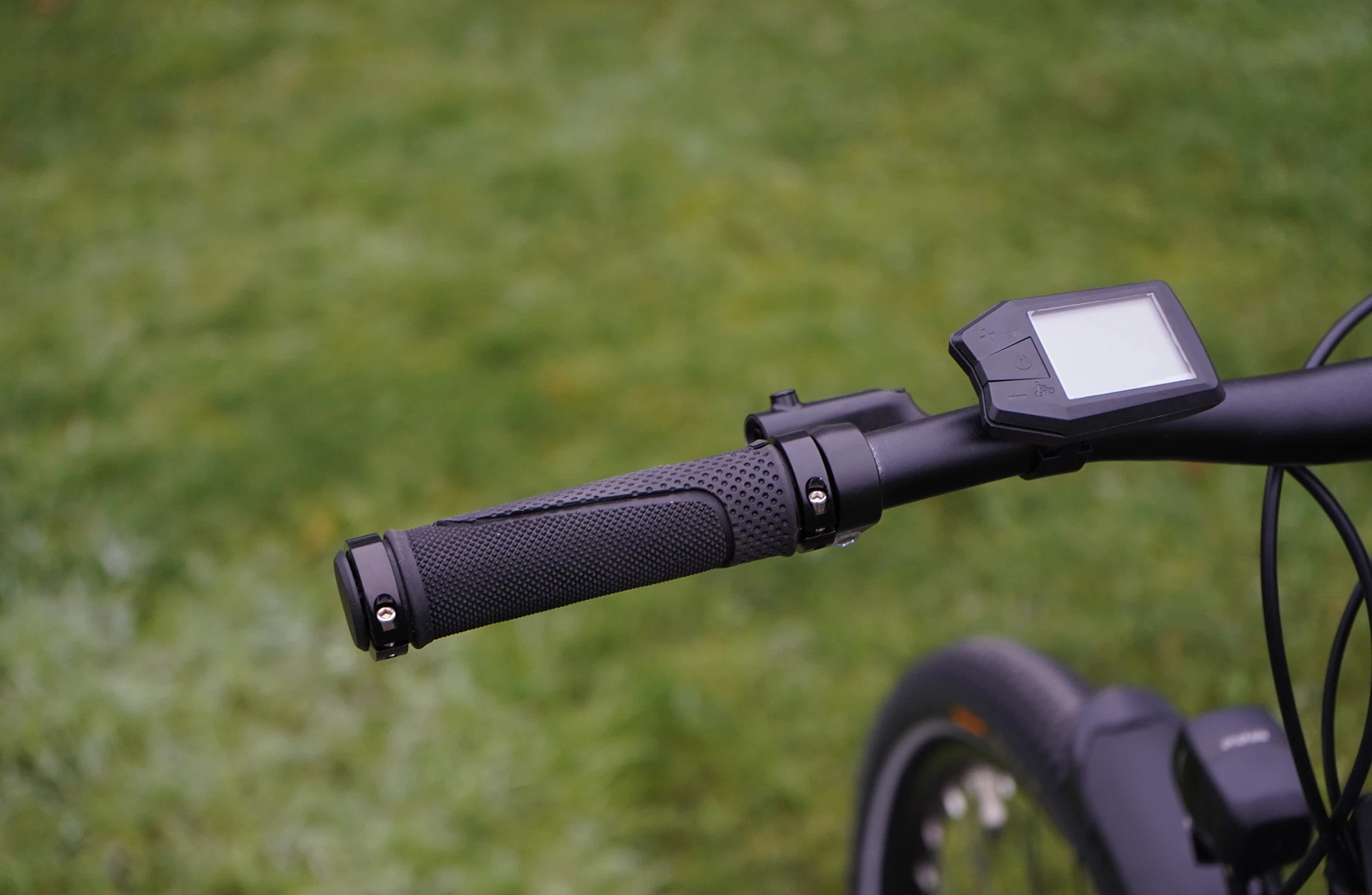 Bildausschnitt linker Lenkergriff mit Bedieneinheit am LCD-Display unscharfem Vorderrad und Scheinwerfer auf Rasen Untergrund
