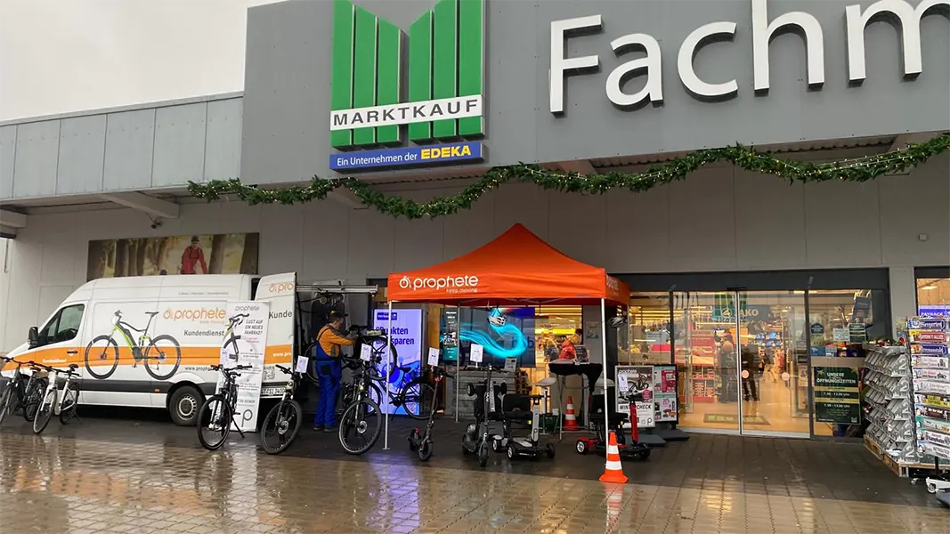 Prophete Wintercheck-Stand mit orangefarbenem Pavillon vor einem Marktkauf, Servicefahrzeug mit Prophete-Branding, E-Bikes und SM4 Kompakt Elektromobil ausgestellt, Infotafeln und Wintercheck-Plakate.