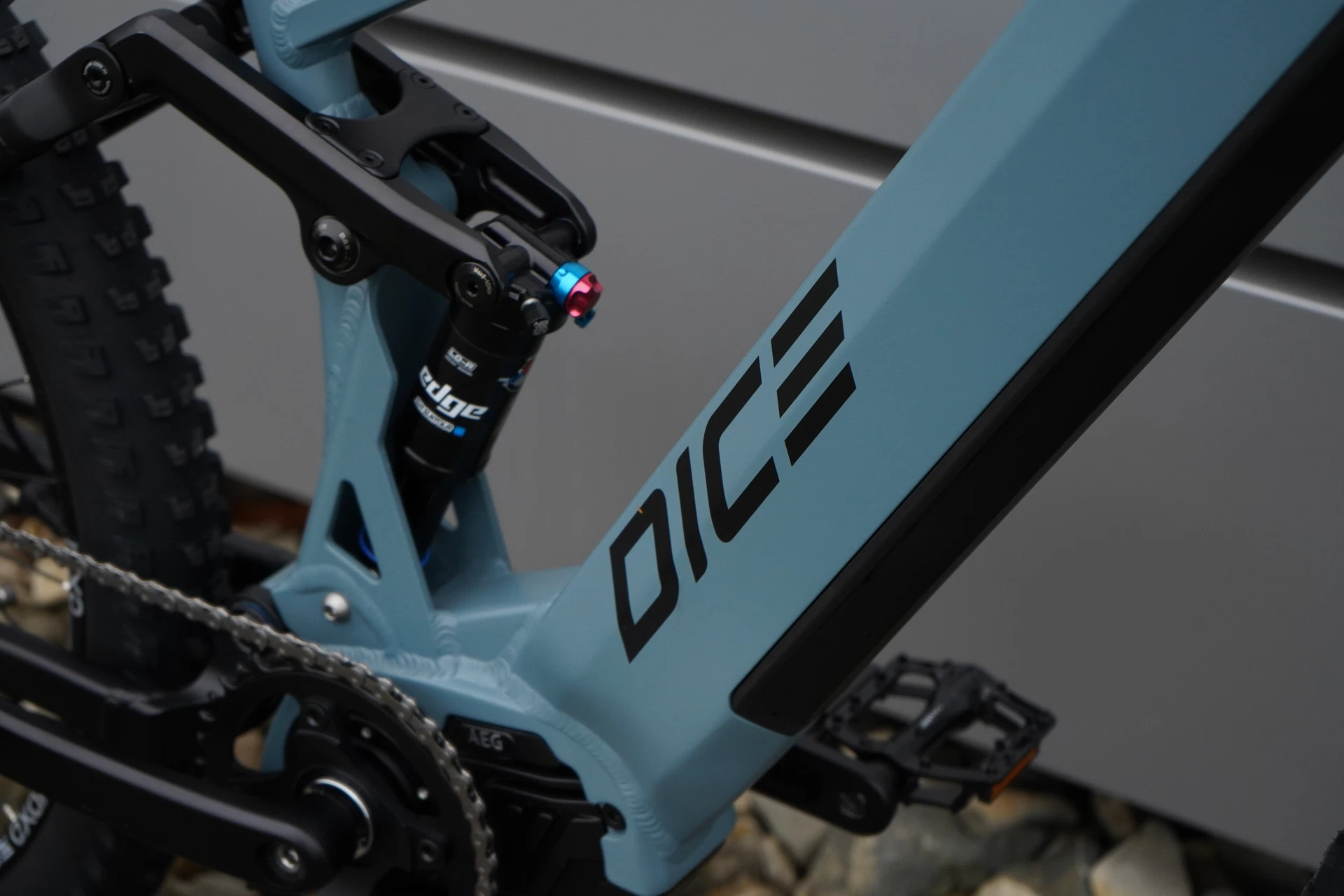 Bildausschnitt Hinterbaudämpfer und DICE Logo am Unterrohr mit vollintegriertem Akku vor Metallwand und auf Steinigem Untergrund