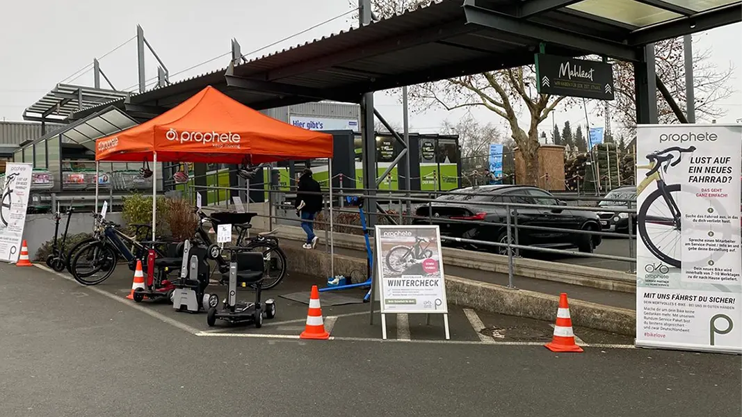 Prophete Wintercheck-Stand mit orangefarbenem Pavillon auf einem Parkplatz, E-Bikes und SM4 Kompakt Elektromobil ausgestellt, Infotafeln und Wintercheck-Plakate vor einem Markt.