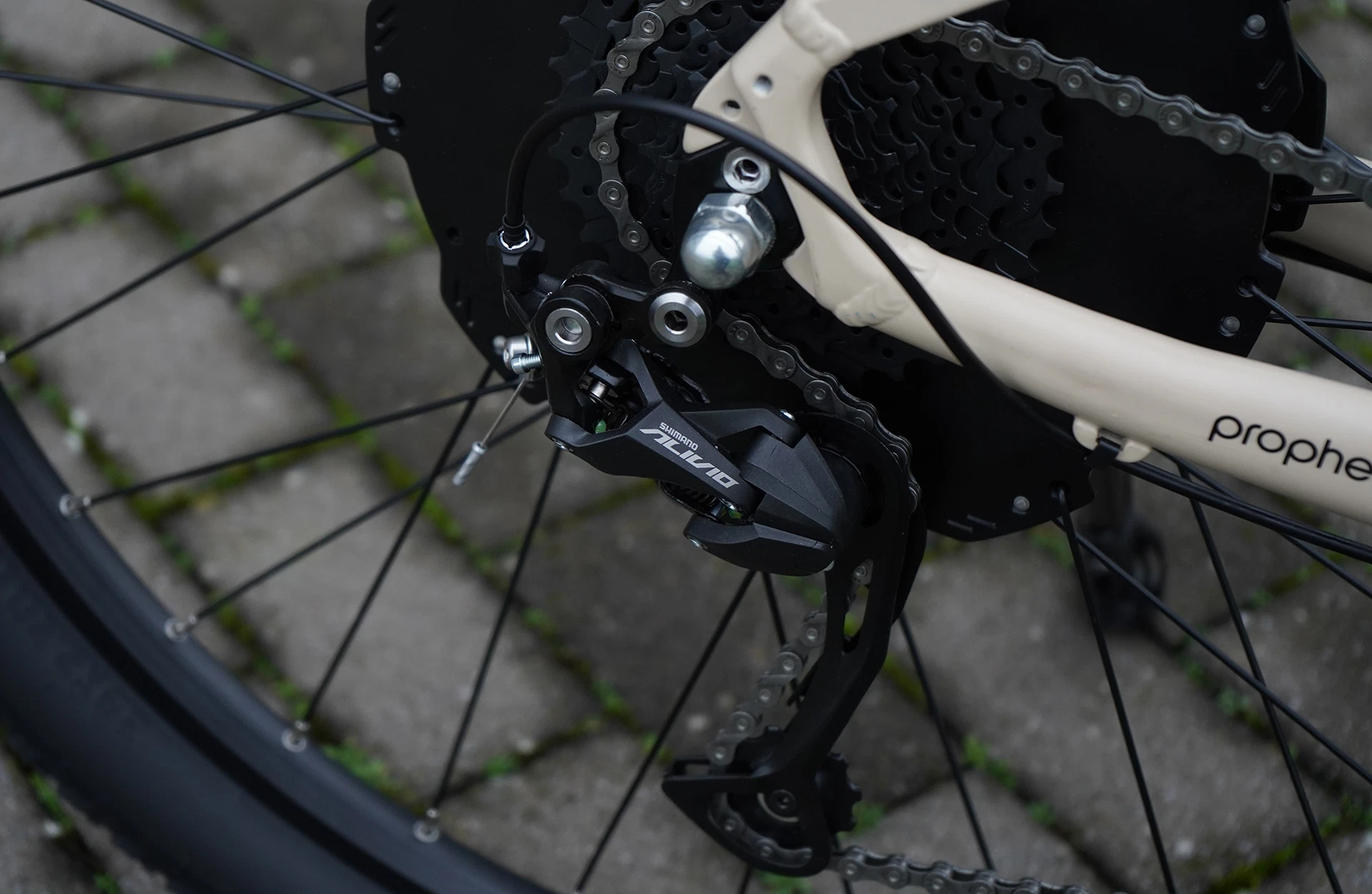 Schwarzes Shimano Alivio schaltwerk an beigem fahrradrahmen, Fokus auf Kettenschaltung und Speichen, gepflasterter Boden im Hintergrund