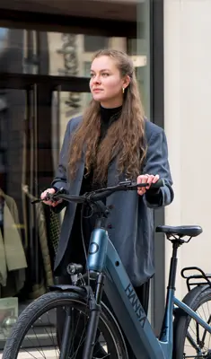 Eine Frau steht im urbanen Umfeld und hält ein modernes Hansa E-Bike an einem Lenker, im Hintergrund eine Glasfassade.
