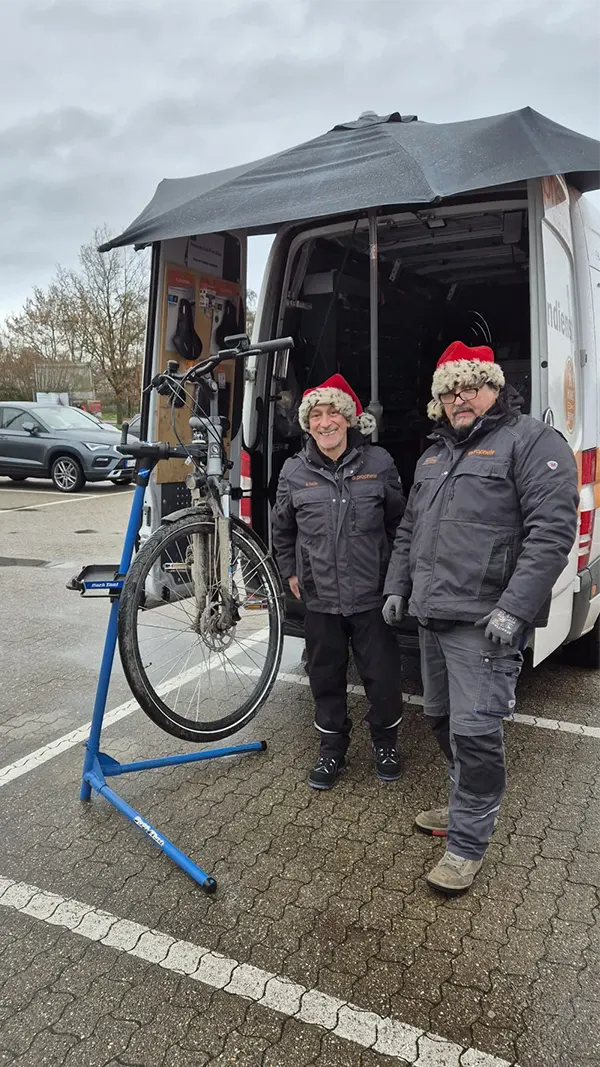 Prophete Wintercheck-Service mit zwei Mitarbeitern in Arbeitskleidung und Weihnachtsmützen, Fahrrad auf Montageständer vor geöffnetem Servicefahrzeug auf einem Parkplatz.