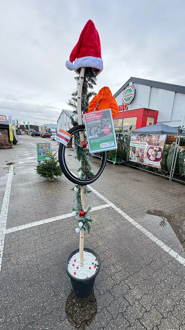 Weihnachtlich dekorierter Fahrradständer mit Santa-Mütze, Prophete-Wintercheck-Poster und Fahrradreifen auf einem Parkplatz vor einem Hagebau.