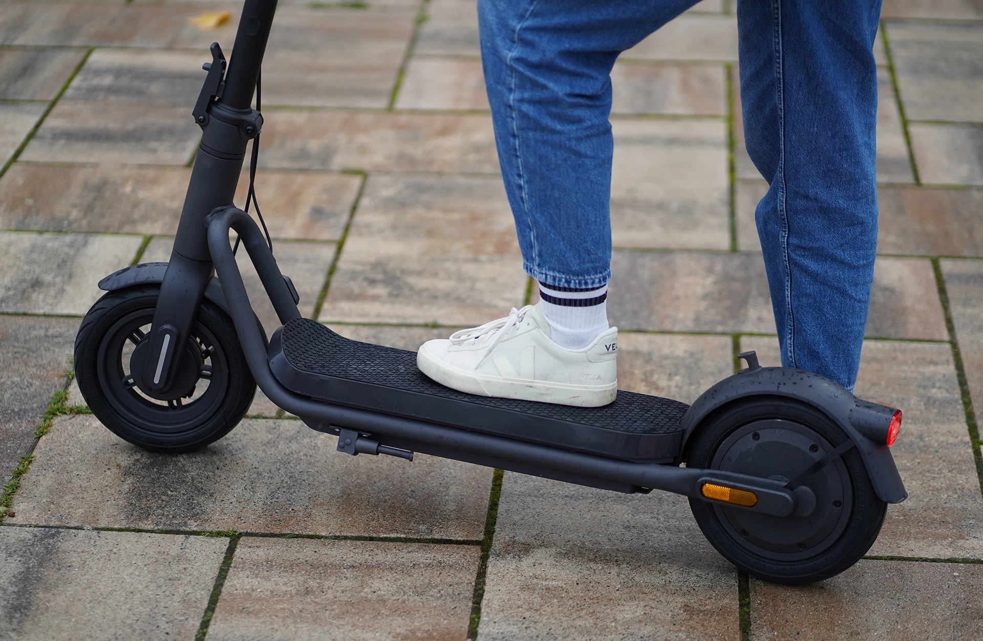 Bildausschnitt unterer Bereich E-Scooter mit Trittbrett und Reifen, mit weißen Sneakers und blauer Jeans vor gepflastertem Untergrund