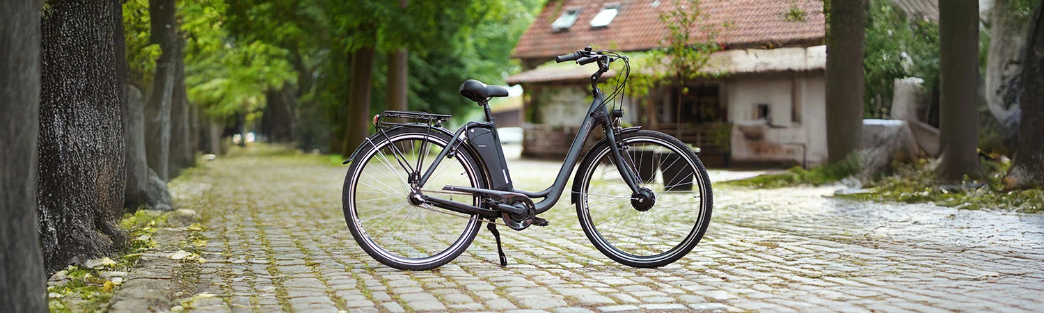 Vollansicht seitlich Prophete Geniesser 1.0 City E-Bike vor dörflicher Kulisse mit Kopfsteinpflaster und Bäumen sowie einem alten Haus
