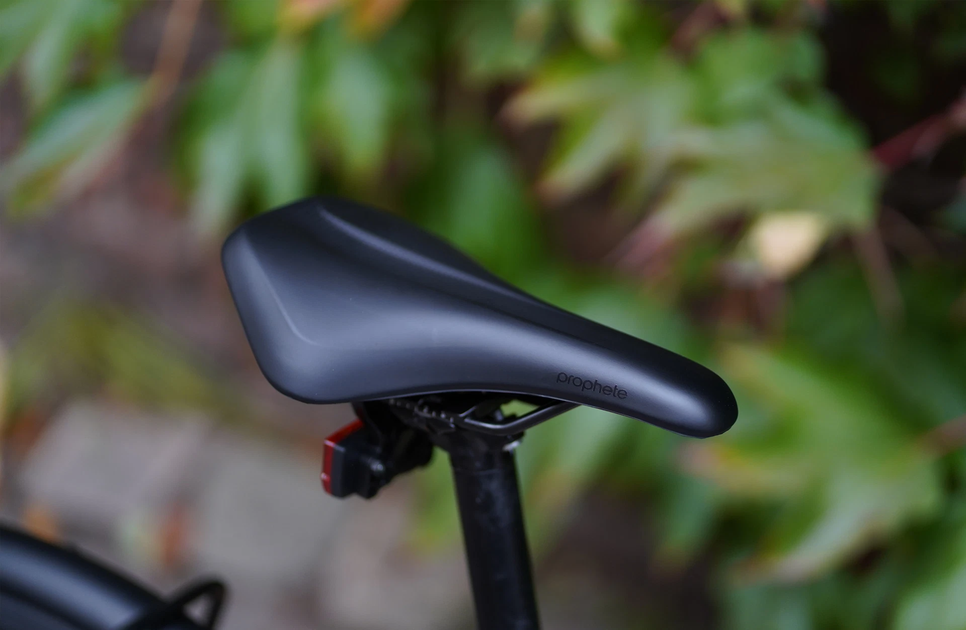 Bildausschnitt Ergonomischer Prophete Sattel mit Logo und angehängtem Reflektor vor blättrigem Hintergrund